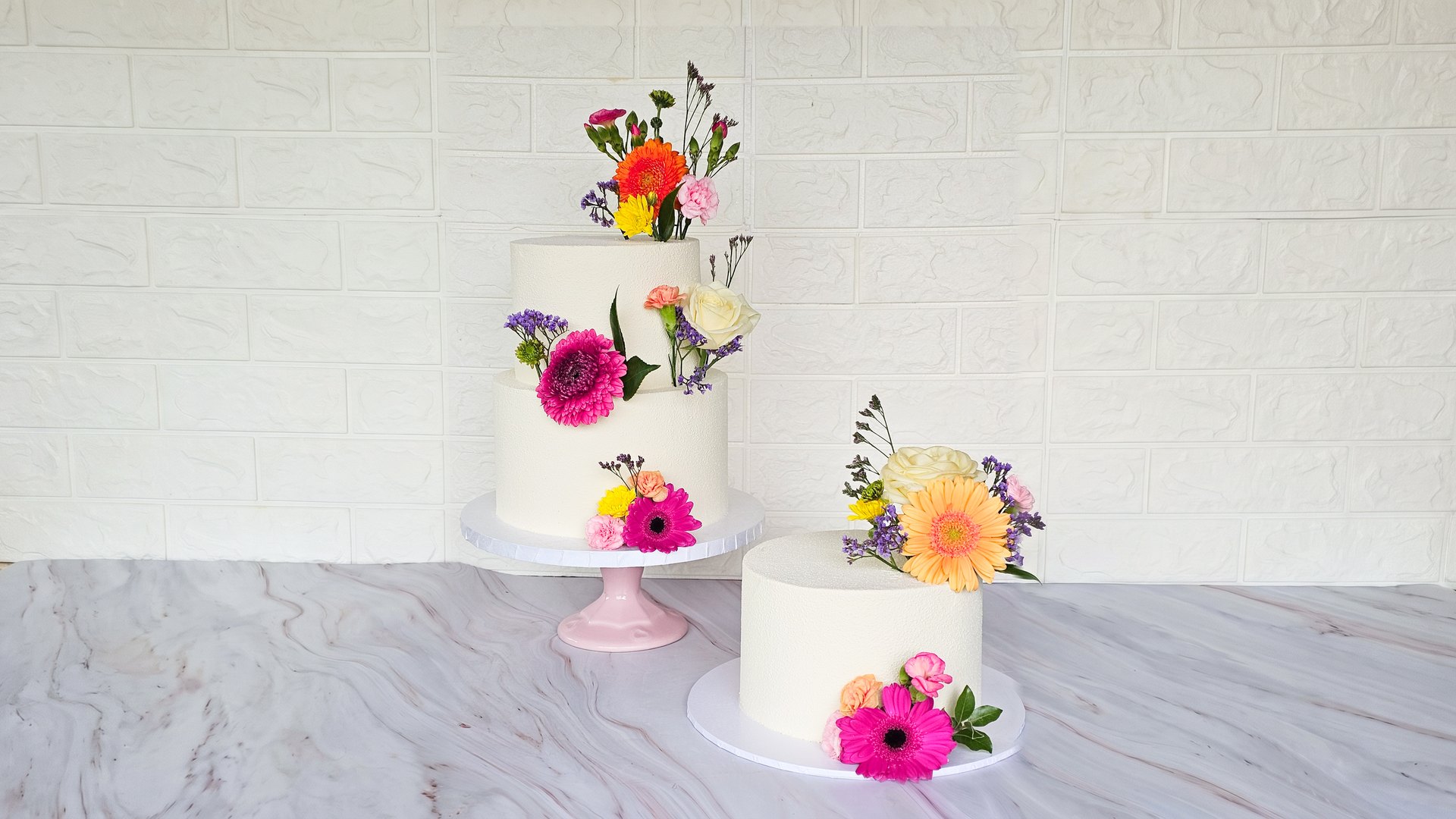Two white cakes decorated with fresh gerberas, carnations and roses on a marble counter, made by Juliana